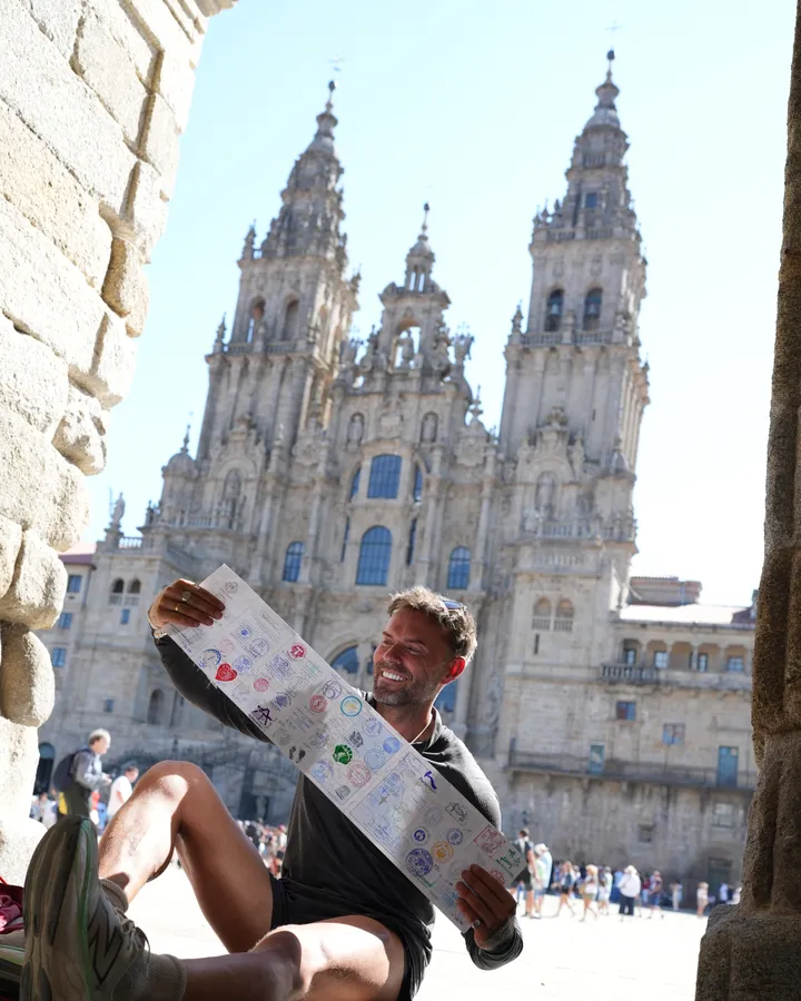 Adrian sitting in the sun outside Santiago de Compostela cathedral, holding up a map and smiling.